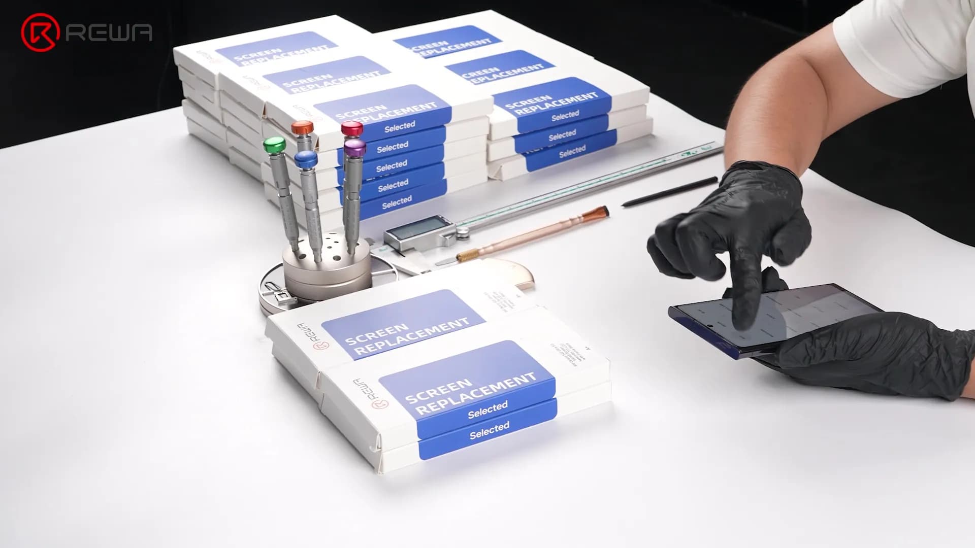 Technician testing a smartphone screen next to stacks of REWA screen replacement boxes and repair tools